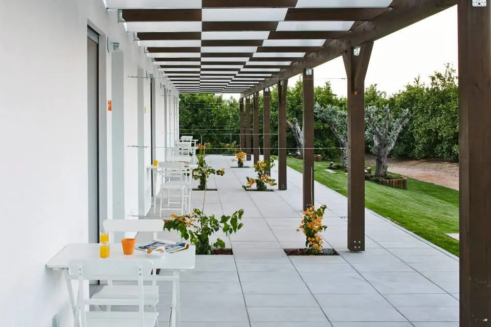 Pérgola de madeira sobre caminho pavimentado, com buganvílias e oliveiras